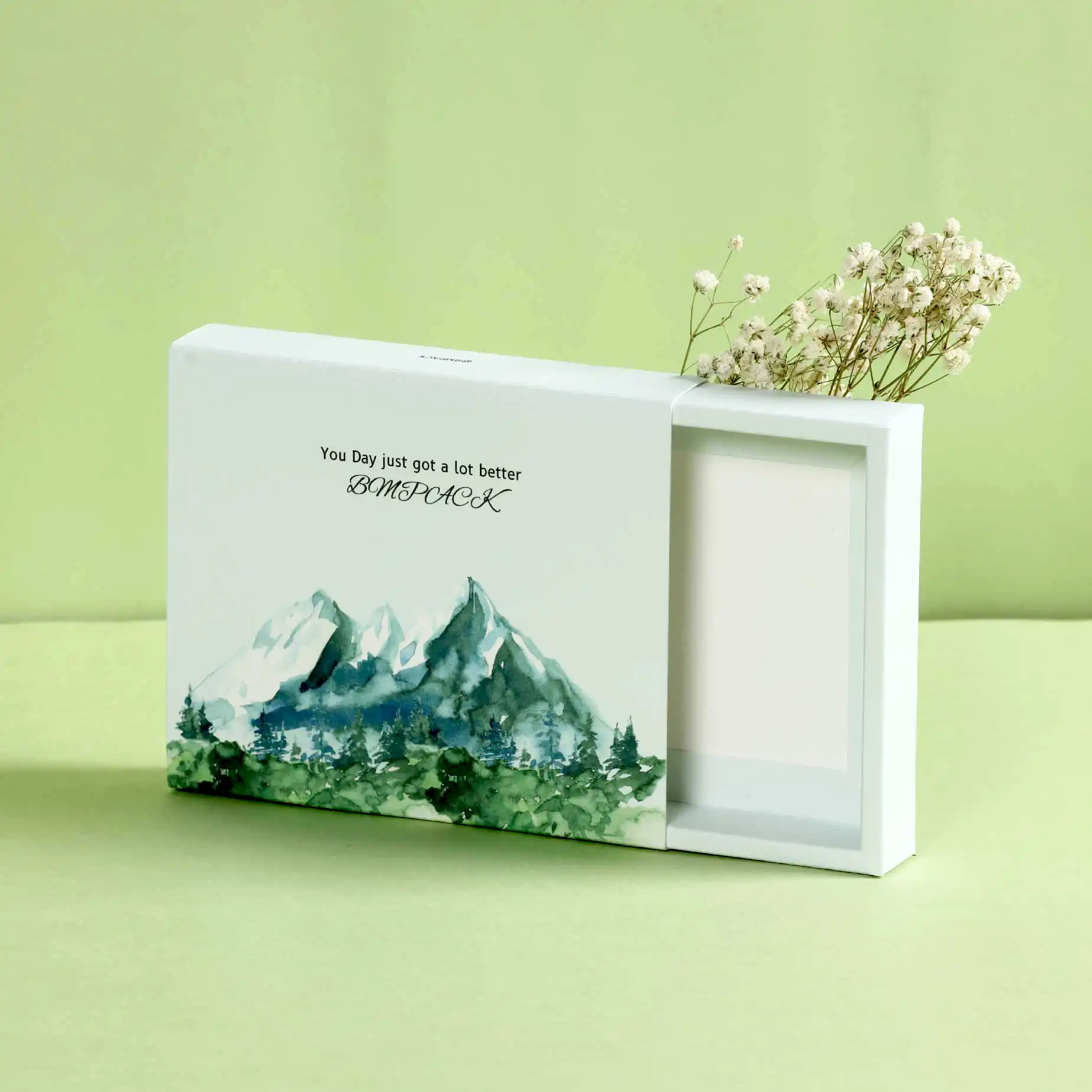 在淺綠色的背景下，擺放了一個湖水綠色的火柴摺盒，盒面印有綠色的森林和樹木，拉出淺綠色內盒，盒旁附有白色乾花。
