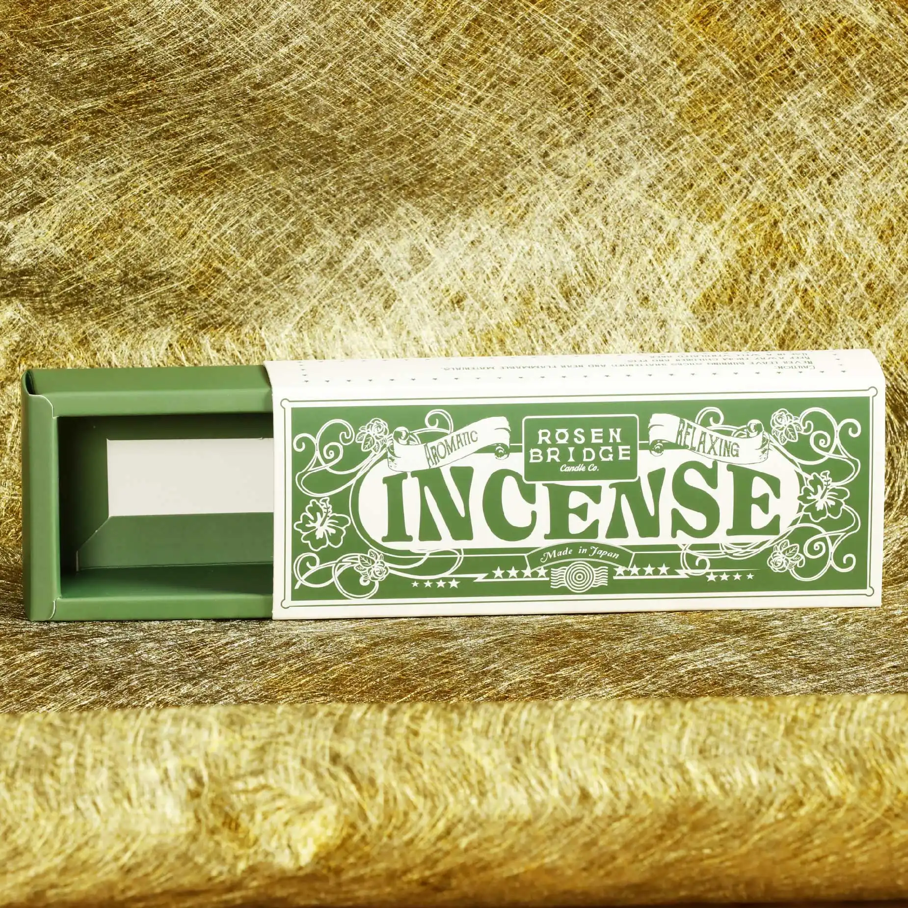 在黃黃綠綠交錯的背景下，擺放了一個白色火柴摺盒，盒面印有"Incense"的字眼和藤本植物的圖像，拉出綠色的內盒。