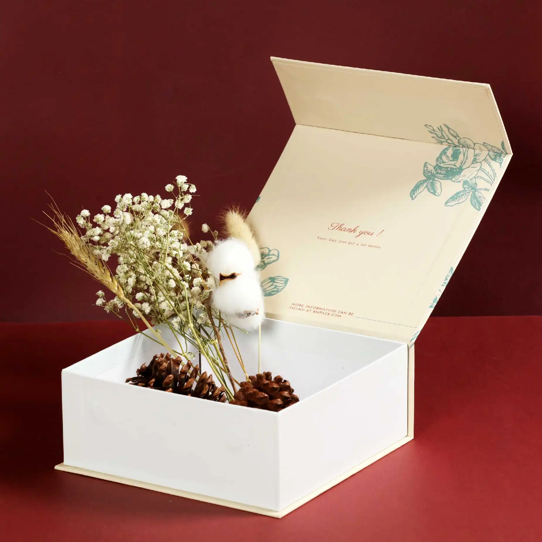 在深紅色的背景下，一個打開了蓋子的磁石書型硬盒，蓋內印有淡黃底色及淺綠色植物圖案。盒內放有五枝乾花和兩顆松果。