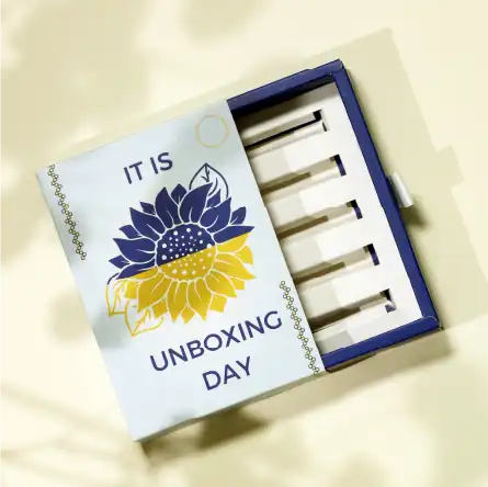 一個白色的抽屜摺盒，盒面印 "IT IS UNBOXING DAY" 及一半黃色和藍色的太陽花圖案，拉出了藍色內盒，盒內有五個產品位置，盒側有一條白色絲帶。
